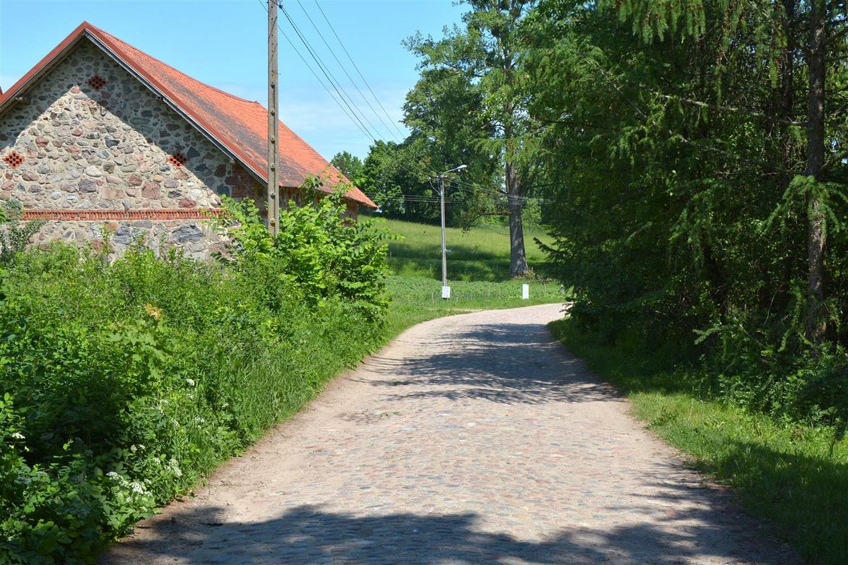 Wąska brukowana droga prowadzi w stronę pola i drzew. Po lewej stronie stoi budynek z kamieni i czerwonym dachem, a wokół rośnie bujna zieleń. W tle widać słup elektryczny i latarnię.