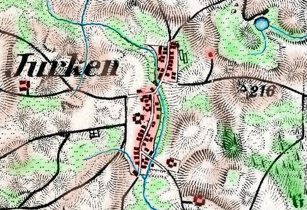 Kolorowa mapa pokazująca wieś Jurken. Na mapie widoczna jest długa ulica z zabudowaniami po obu stronach zaznaczonymi na czerwono. Otaczają ją pola, wzgórza i lasy w kolorach zielonym i beżowym. Przez miejscowość przepływa cienka niebieska linia oznaczająca strumień lub małą rzekę. W pobliżu zaznaczono wysokość terenu – 216 metrów. Mapa ma styl topograficzny.