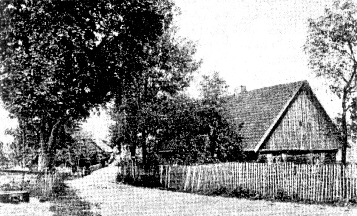 Czarno-biała fotografia przedstawia wiejską drogę otoczoną drzewami. Po prawej stronie widać drewniany dom z dwuspadowym dachem i ogrodzeniem z drewnianych sztachet. W tle widać więcej domów i drzewa rosnące po obu stronach drogi. Zdjęcie ukazuje spokojną, wiejską scenerię.