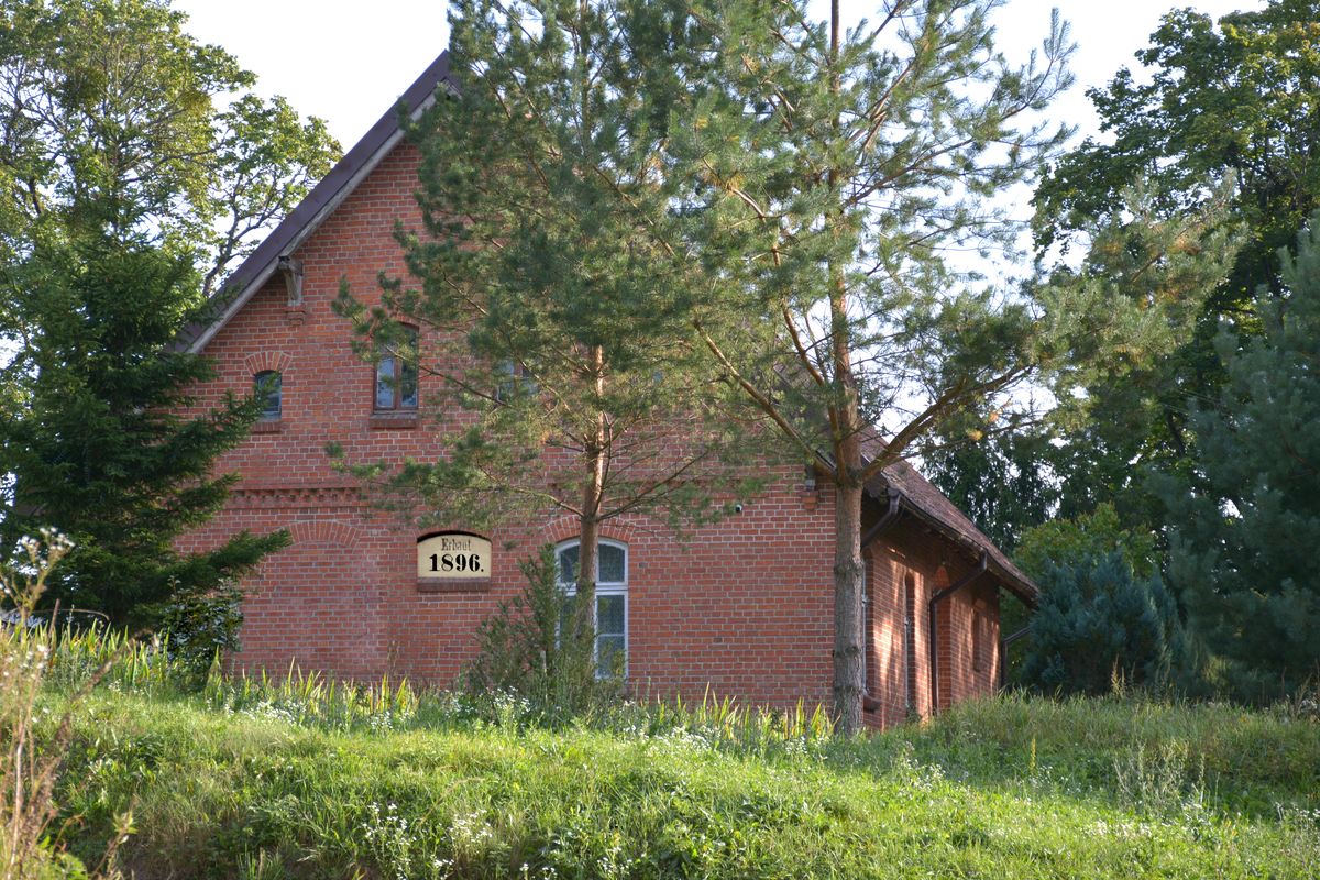 Ceglany dom z 1896 roku częściowo zasłonięty drzewami, stoi na trawie wśród zieleni.