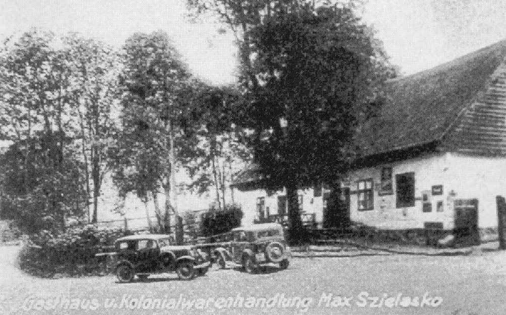Czarno-biała fotografia przedstawiająca dawny budynek z szyldem „Gasthaus u. Kolonialwarenhandlung Max Szielasko”, czyli gospoda i sklep kolonialny Maxa Szielasko. Przed budynkiem stoją dwa stare samochody. W tle widać wysokie drzewa, a sam budynek ma spadzisty dach i białe ściany z ciemnymi oknami i drzwiami.