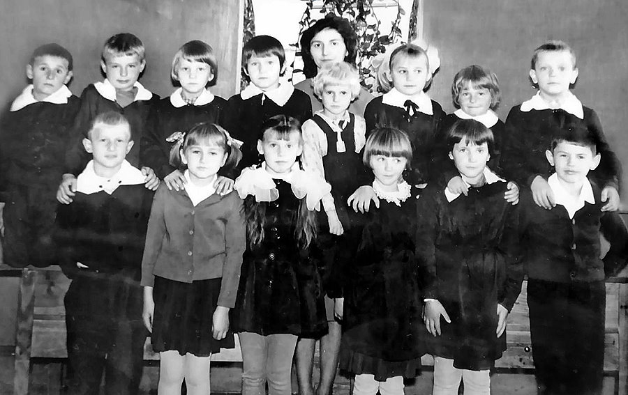 Czarno-białe zdjęcie grupowe przedstawiające klasę szkolną z lat 60. lub 70. XX wieku. Na fotografii widać piętnaścioro dzieci w szkolnych mundurkach – dziewczynki w sukienkach z białymi kołnierzykami, chłopcy w marynarkach lub swetrach. W środku w górnym rzędzie stoi nauczycielka z ciemnymi włosami. Dzieci stoją w trzech rzędach, uśmiechają się lekko lub patrzą poważnie w stronę aparatu. Zdjęcie wykonano w klasie – w tle widać ścianę i roślinę w doniczce.