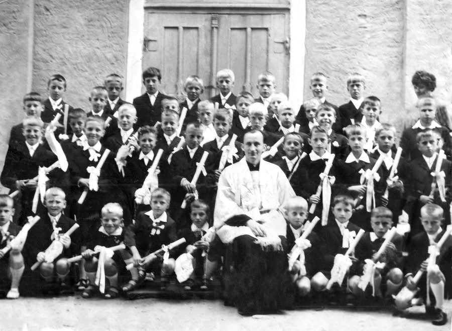 Czarno-białe zdjęcie grupowe przedstawiające dużą grupę chłopców ubranych w eleganckie garnitury z białymi kokardami, trzymających świece — prawdopodobnie podczas uroczystości pierwszej komunii świętej. W środku siedzi ksiądz w komży, otoczony przez dzieci. Zdjęcie wykonano przed kościołem, widoczne są drzwi i tynkowana ściana.