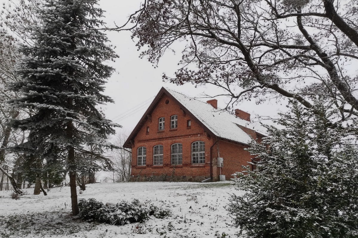 Ceglany budynek z dużymi oknami i dwuspadowym dachem pokrytym śniegiem, otoczony drzewami i krzewami zimą. Na ziemi leży cienka warstwa śniegu, a drzewa mają lekko ośnieżone gałęzie. Scena jest spokojna i zimowa.