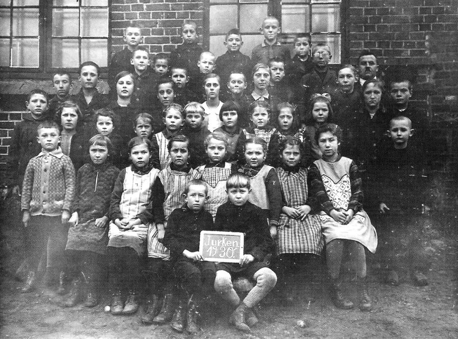Czarno-białe zdjęcie grupowe dzieci i nauczyciela przed szkołą. Na tabliczce widnieje napis „Jurken 1930”
